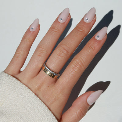 A hand showcasing salon-quality, long, almond-shaped Laura Press-On Nails in baby pink by Glowie By Her, each nail adorned with a small rhinestone near the cuticle. The hand is accessorized with two gold rings: one simple band and one featuring alternating patterns. A sleeve of a light-colored garment is also visible at the wrist.