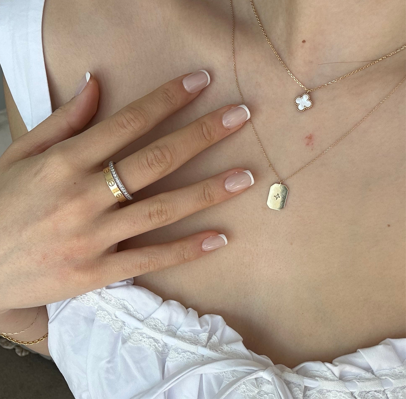 A person wearing a white top showcases a delicate necklace with a flower pendant and another one with an engraved medal. Their nails have the salon-quality French tip look, thanks to Christi Press-On Nails by Glowie By Her, and they wear a gold ring adorned with small diamonds on their ring finger.