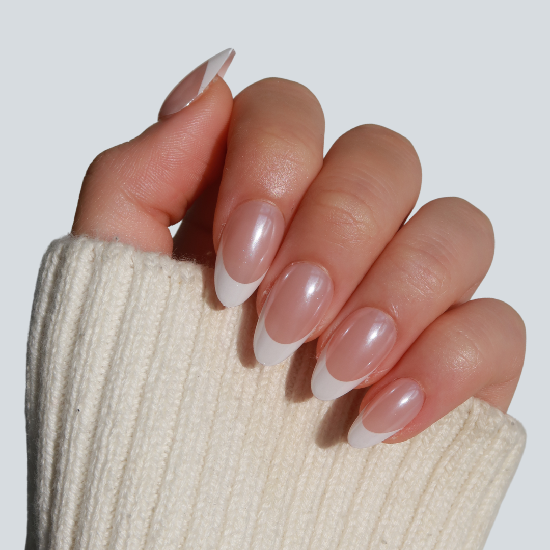 A hand with a French manicure of Femme Fatale natural pink press-on nails with white tips from Glowie By Her is against a light background. Partially covered by a cream-colored sweater sleeve, it showcases vegan, cruelty-free elegance.