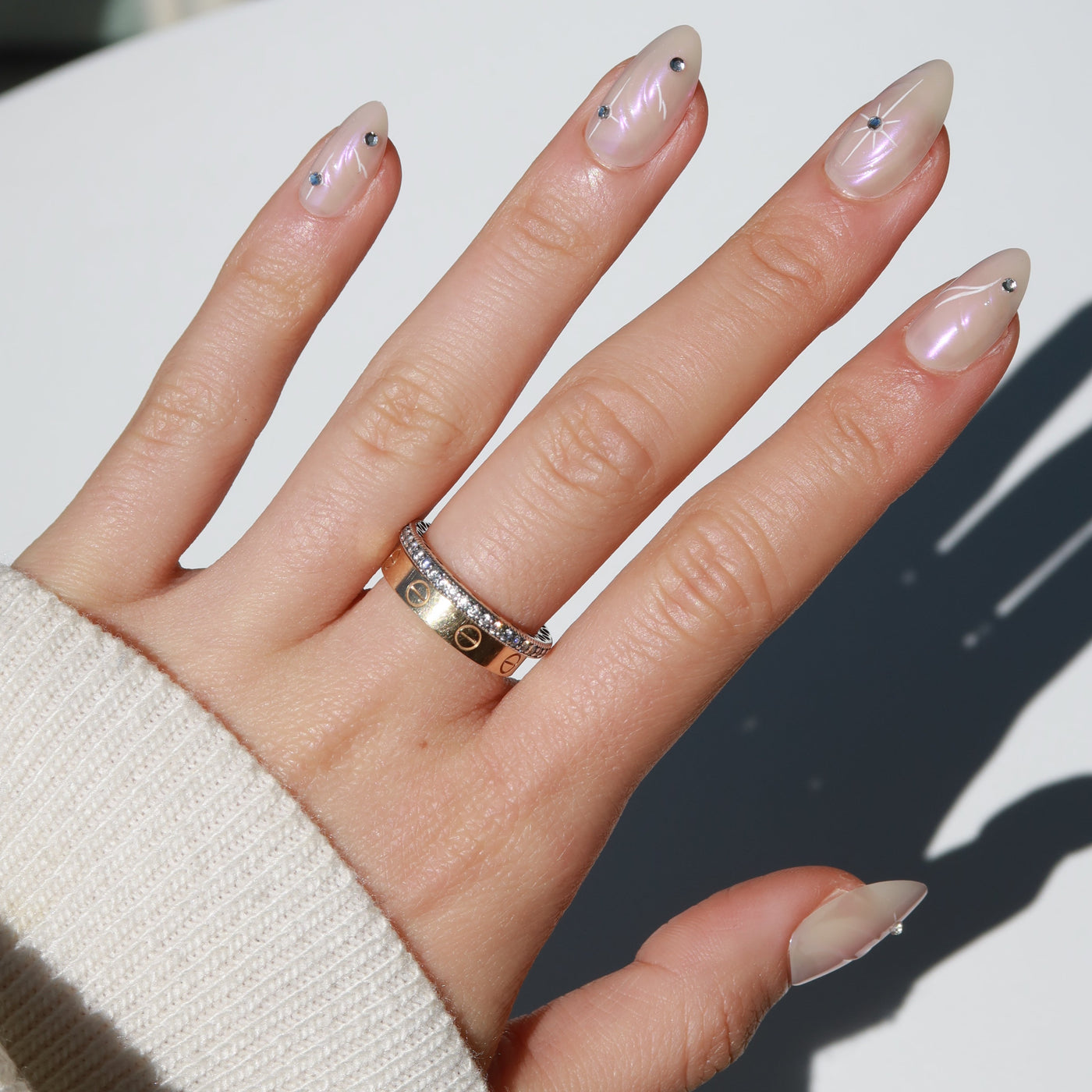 A hand with a beige sweater sleeve showcases manicured, glossy almond nails adorned with small decorative dots and stars. The hand is graced with two rings: one gold with small cutout circles and another, above it, featuring a row of diamonds. The polished look is achieved using the Aura Press-On Nails from Glowie By Her.