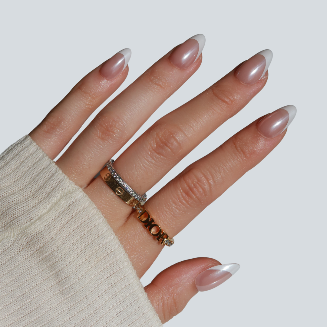 A hand with long, almond-shaped nails showcases Glowie By Her's Femme Fatale, featuring a chic chrome French tip gradient from translucent pink to white tips. The fingers wear two gold rings and one gemstone-encrusted silver ring, paired with a beige sweater.