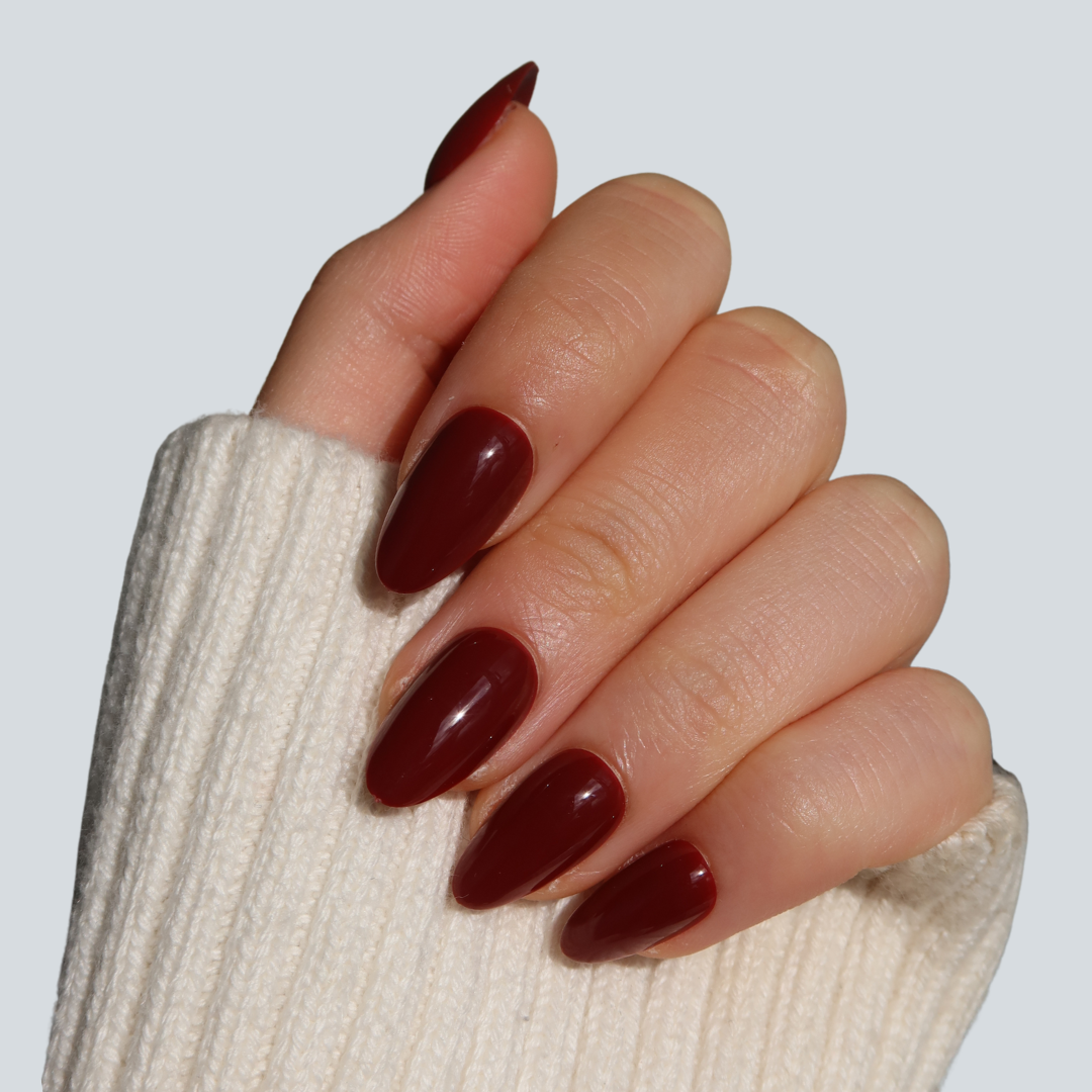 A hand with long, oval nails showcases Glowie By Her's "Smitten," a glossy dark red polish akin to medium cherry cola. Partially covered by a cream knit sweater sleeve, it contrasts against a plain light gray background.