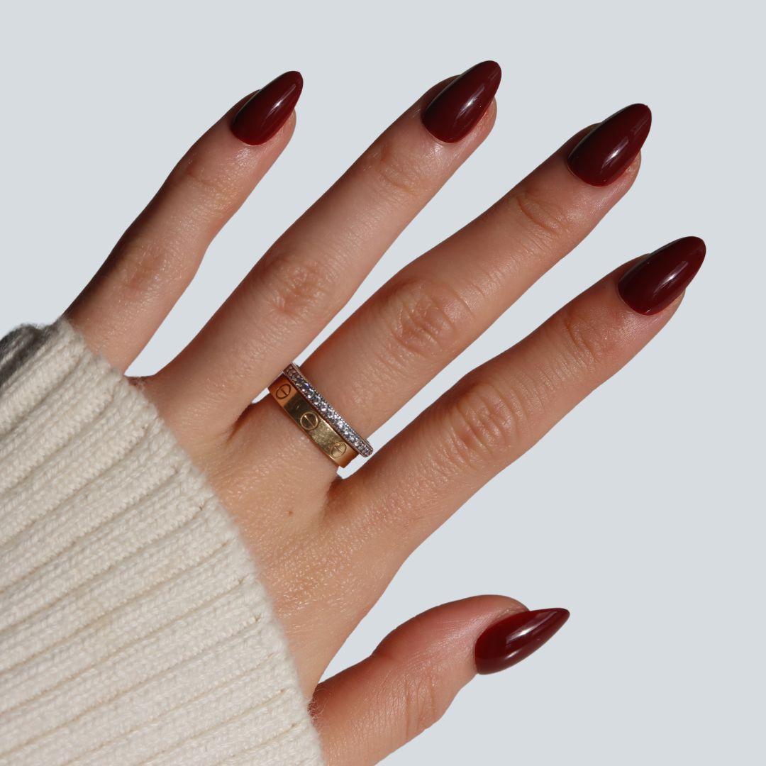 A hand with Smitten medium cherry cola nails by Glowie By Her wears a gold and a silver ring with small stones, paired with a cream ribbed sweater. The light gray background highlights the salon-quality nails' striking color and elegant design.