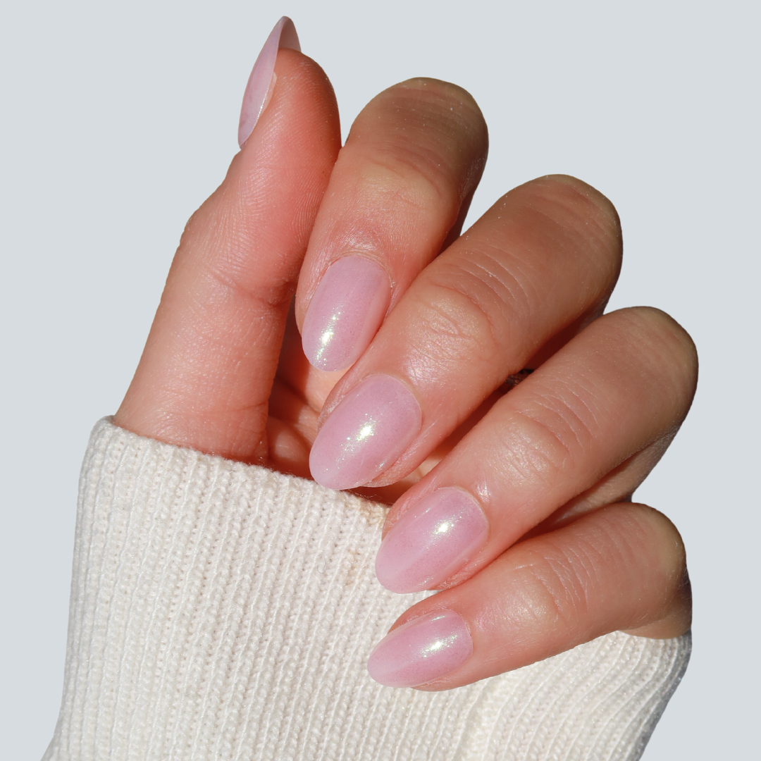 A hand adorned with Glowie By Her's Fairydust Press-On Nails, featuring neatly manicured, almond-shaped nails polished in a glossy light pink shade, is shown. The person is wearing a white knit sweater against a simple solid light gray background. These salon-quality nails boast an elegant iridescent chrome finish that adds a touch of sophistication.