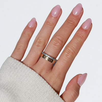 A hand adorned with Glowie By Her's Fairydust Press-On Nails, featuring salon-quality, light pink, oval-shaped nails, is wearing two stacked rings on the middle finger. The top ring is a gold band encrusted with small diamonds, while the bottom ring is a wider gold band with intricate engravings. The person is also wearing a cream-colored sweater.