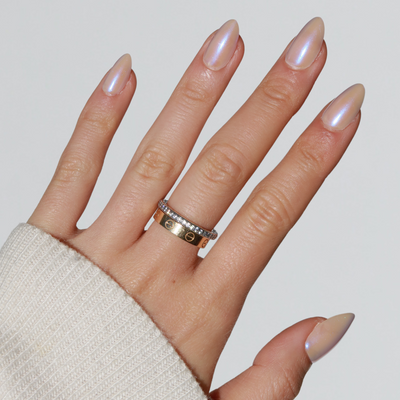 A hand showcasing salon-quality nails using 777 Press-On Nails by Glowie By Her is adorned with two rings on the ring finger. One ring features a simple gold band with a repeating pattern, while the other is a limited edition silver band set with small diamonds. The hand is partially covered by the sleeve of a light-colored sweater.