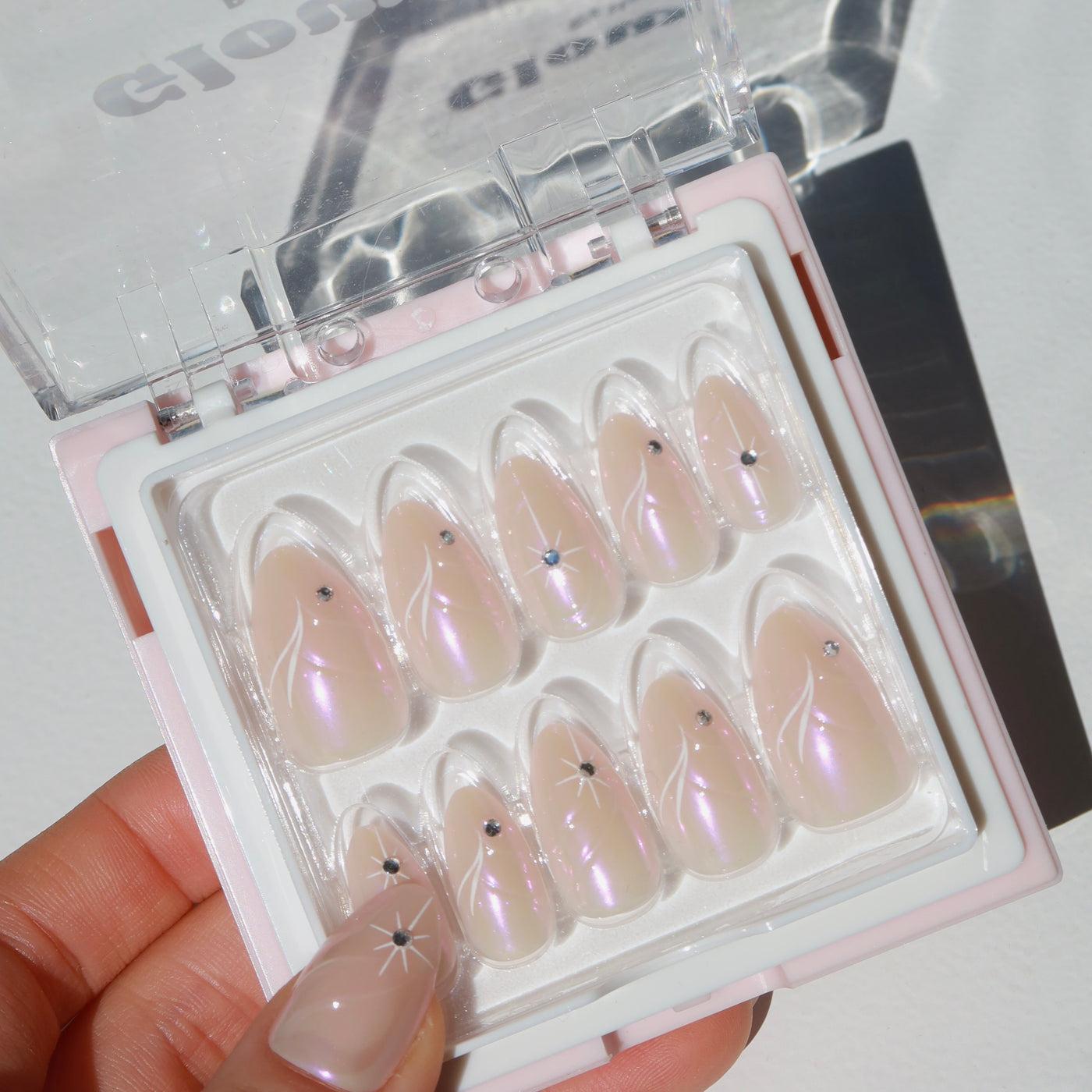 A hand with manicured nails, featuring a star design, holding an Aura Press-On Nails kit by Glowie By Her. The nails in the open box are glossy almond shapes in light pink or white, some adorned with iridescent pink chrome accents and small star-shaped designs with gem details. The arrangement is neat and appealing.