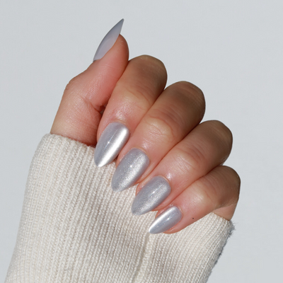 A hand with a light skin tone is showcased in a cream sweater, flaunting salon-quality, almond-shaped nails. The glossy, metallic silver polish from Glowie By Her's "Haze" collection adds a touch of elegance against the plain white background.