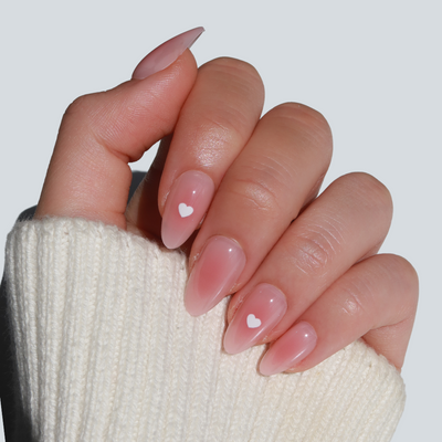 A hand with long, almond-shaped nails painted in Glowie By Her's "Mommy," a salon-quality blushed pink polish. Featuring small white heart designs and 100% vegan & cruelty-free ingredients, the soft tones are beautifully contrasted by a cream-colored knitted sleeve.