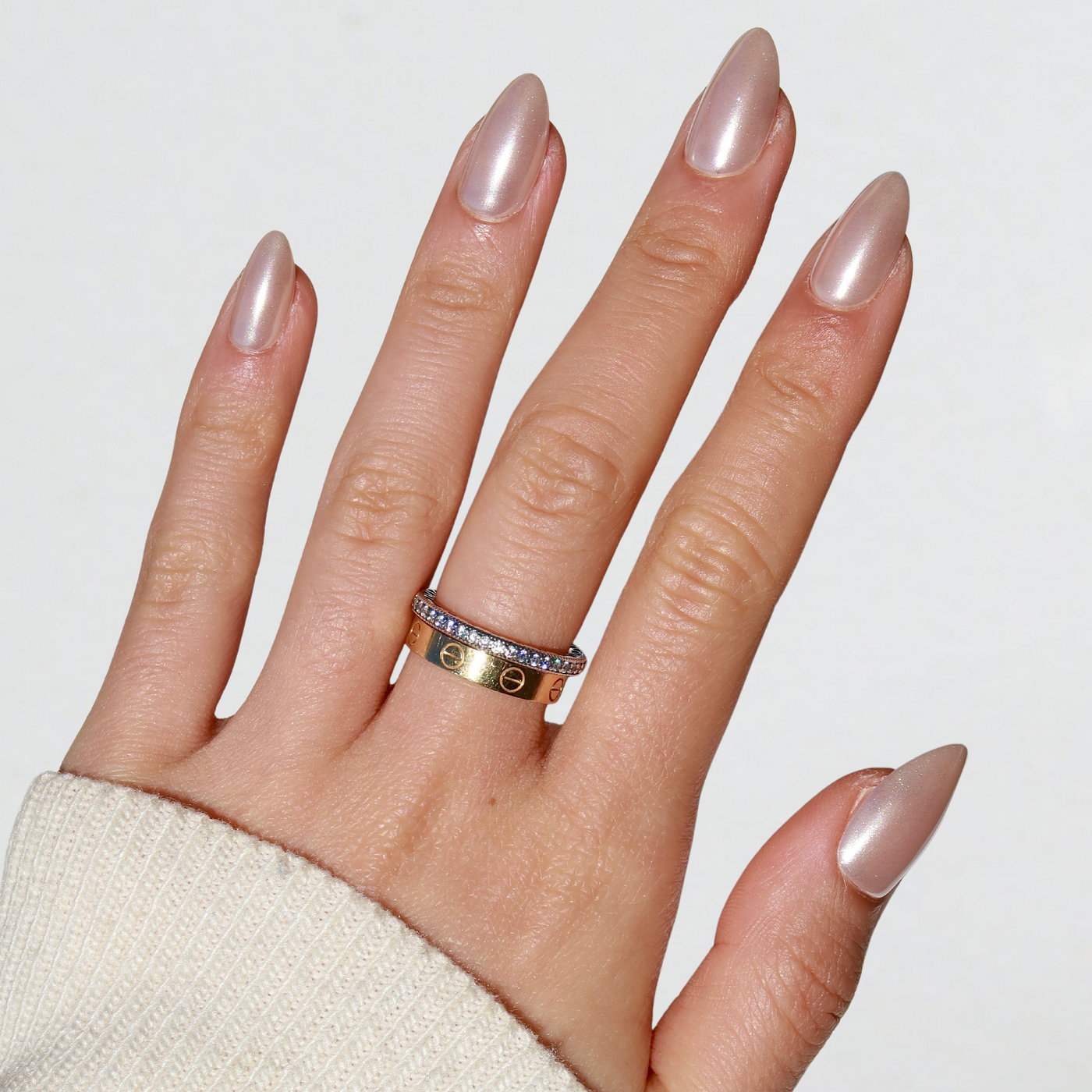 A hand with long, almond-shaped Allure Press-On Nails by Glowie By Her, painted in a metallic nude shade, is shown. The index finger showcases two rings: one gold with engraved letters and another silver band encrusted with small diamonds. These salon-quality nails perfectly complement the light-colored, ribbed sleeve.