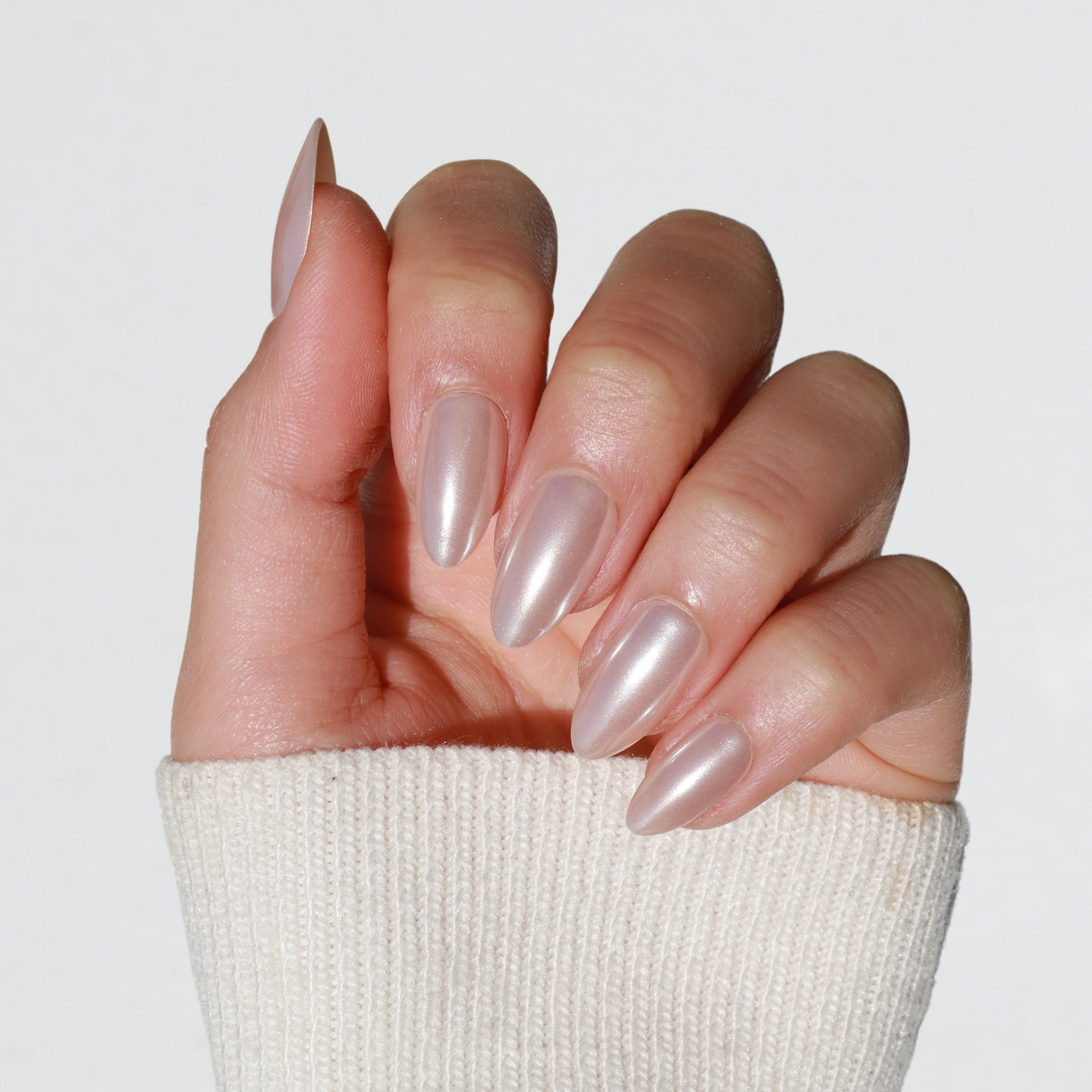 A hand adorned with Glowie By Her's Allure Press-On Nails, featuring long, almond-shaped tips in an iridescent, shimmering nude polish, set against a plain white background. The hand is partially wrapped in the oversized sleeve of a light-colored, textured sweater.