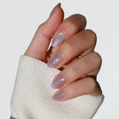 A hand featuring Glowie By Her's Celestial polish, a salon-quality glittery light mauve-pink, is shown with fingers slightly curled against a light background. The person is wearing a cream-colored knit sweater that partially covers the hand.