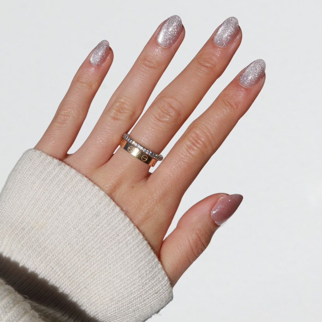 A hand showcasing the Celestial glittery light mauve-pink nails by Glowie By Her is displayed against a white backdrop. The individual is dressed in a cream sweater, and their middle finger is adorned with two gold rings: one simple band and another with small embellishments, creating a salon-quality effect.