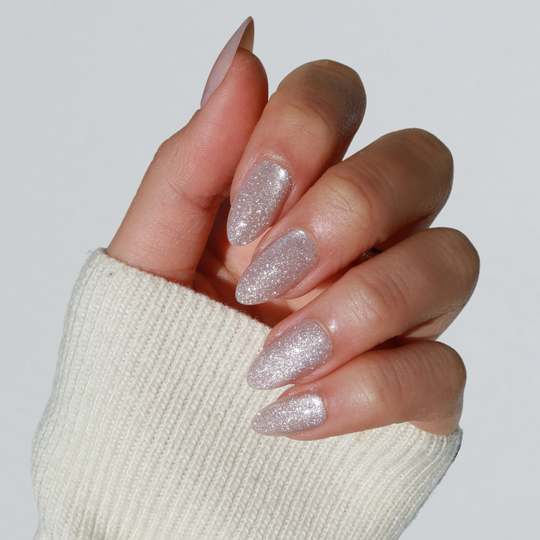 A hand adorned with long, pointed nails showcases the salon-quality shimmer of Glowie By Her's Make a Wish silver glitter polish. The person is wearing a cream-colored ribbed sweater, and the nails are elegantly posed against a plain gray background.