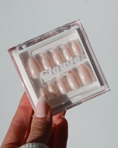 A person with neatly manicured nails holds a clear plastic case containing a set of light pink, glossy press-on nails of varying sizes from the "Sweet Talk Collection" by Glowie By Her. The baby pink nails feature the brand name "Glowie By Her" written on the top and come with optional rhinestone accents for added glamour. Currently, they are available at 20% off.