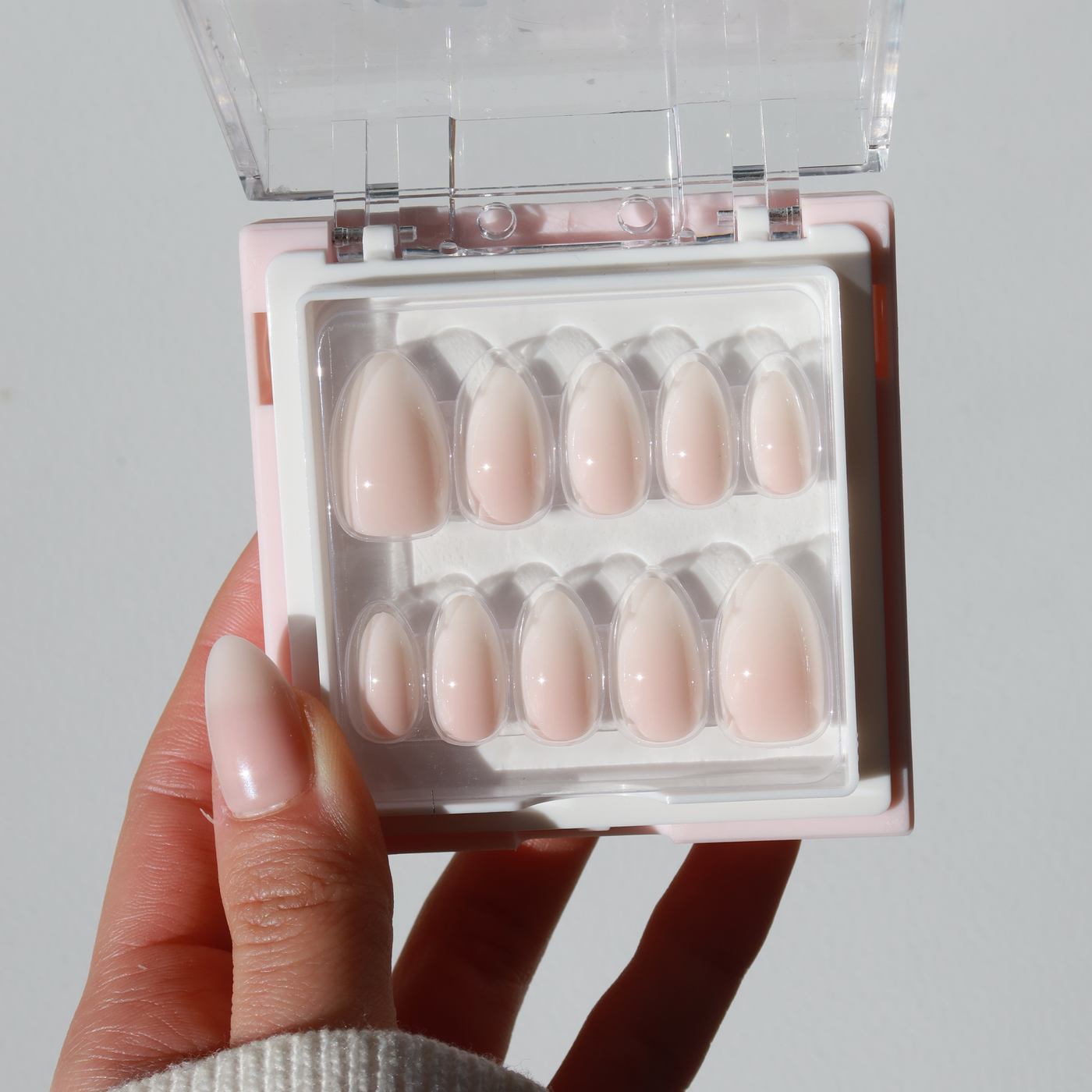 A hand holds a small rectangular box containing a set of salon-quality, pink almond-shaped artificial nails in various sizes. The box is open, revealing ten short nails from the Glowie By Her Daydream Press-On Nails (Short) collection, arranged in two rows on a white tray. The background is a simple, light-colored surface.