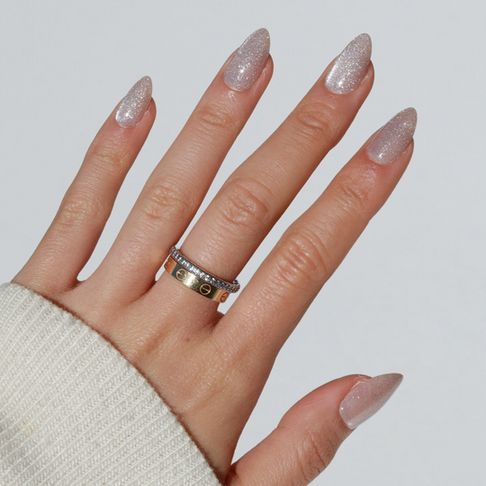 A hand with long, glittery, almond-shaped nails flaunts the "Make a Wish" cat-eye effect from Glowie By Her against a plain background. Adorned with two rings—a gold band engraved with intricate designs and a silver ring set with small stones—the hand emerges from the cozy sleeve of a light-colored sweater, showcasing salon-quality nails.
