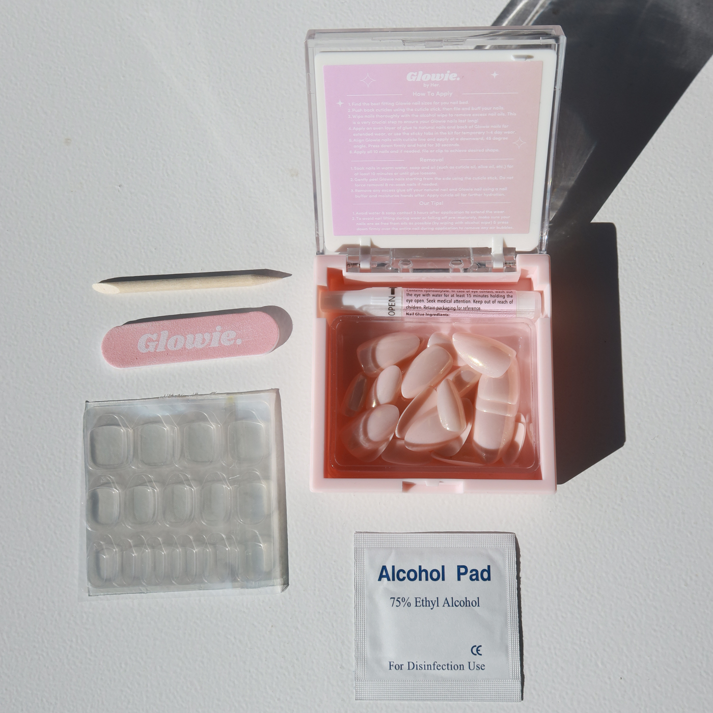 A nail care set featuring "Glowie By Her" Daydream Press-On Nails (Short), a pink emery board labeled "Glowie," a wooden cuticle pusher, a clear blister pack of adhesive tabs, a small pink case of salon-quality nails in short almond style, and one alcohol pad packet.