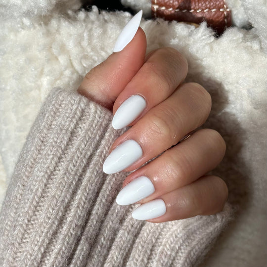 A hand with salon-quality, almond-shaped nails painted in Glowie Co.'s Snow White peeks out of a cozy beige knitted sleeve against a soft, fluffy white fabric background.