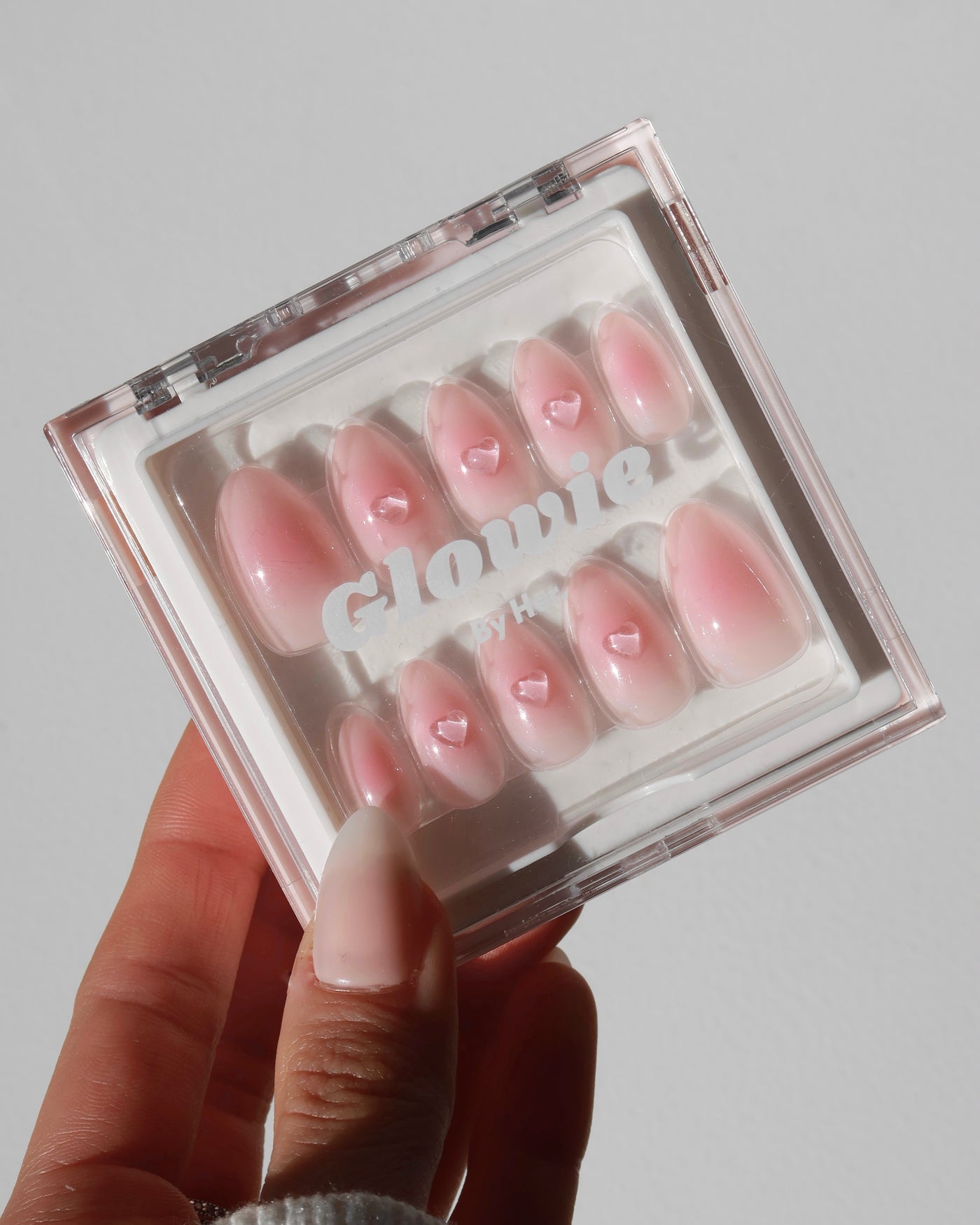 A hand holds a square transparent box containing a set of baby pink press-on nails from the Sweet Talk Collection, which is currently 20% off. Each nail features a small heart design near the tip and rhinestone accents, topped with a glossy finish. The box displays the text "Glowie By Her" on its front.