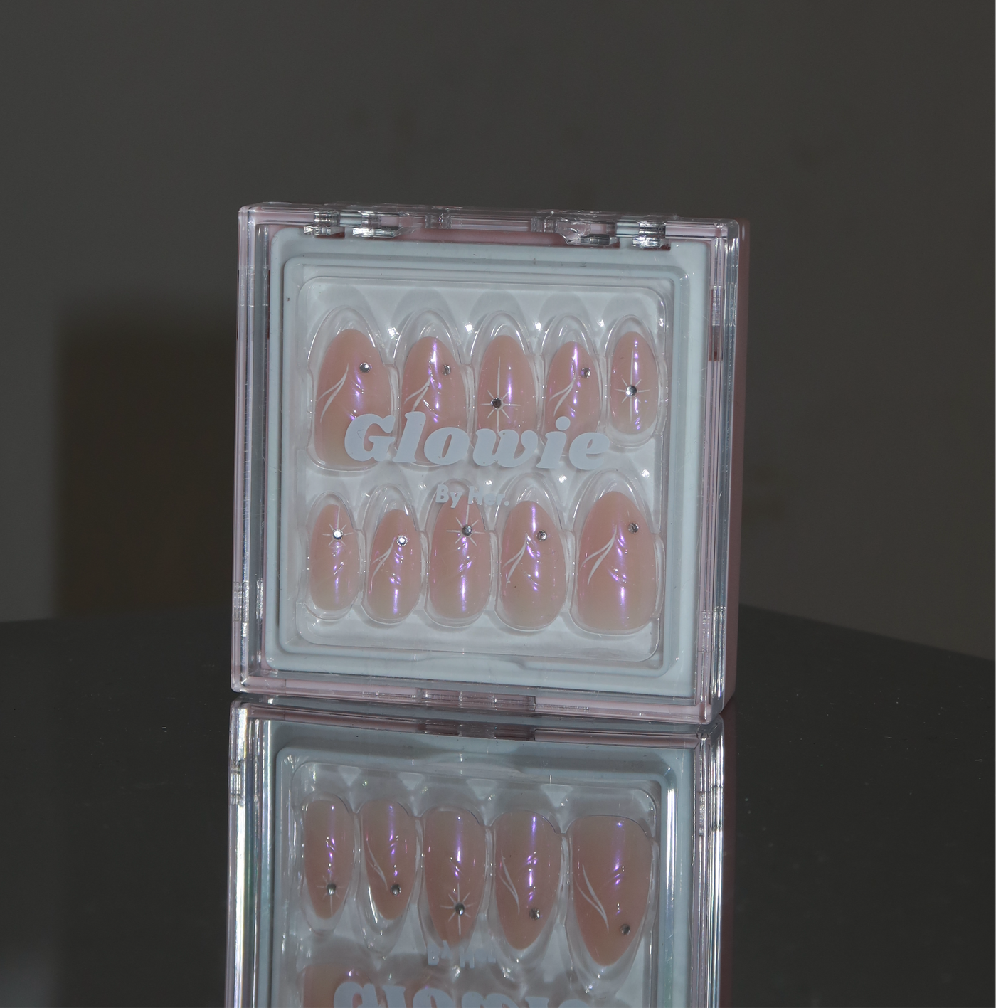 A clear box labeled "Glowie By Her" displays a set of iridescent pink chrome Aura Press-On Nails with tiny black star decorations inside. The glossy almond nails are showcased on a reflective surface, casting a partial mirror image below.