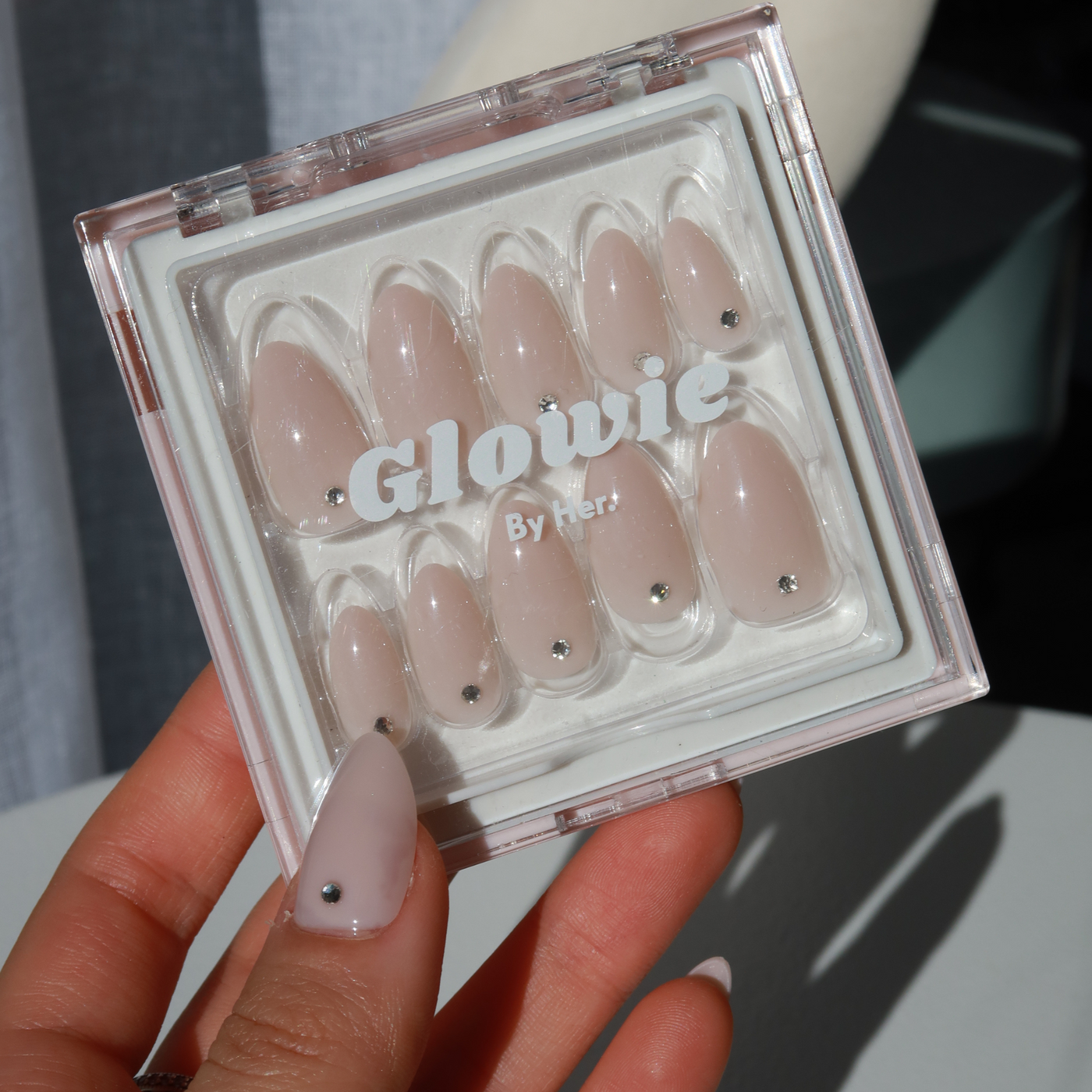 A hand with light-colored, almond-shaped nails holds a transparent box labeled "Glowie By Her," containing a set of Laura Press-On Nails. Each baby pink nail is adorned with a small, round, silver embellishment near the cuticle.