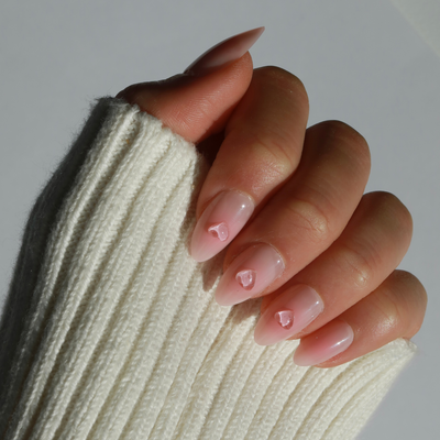 A hand with a cream-colored knitted sleeve partially covering the fingers showcases almond-shaped nails adorned with Made You Blush Press-On Nails by Glowie By Her. These nails feature a blush base and are accented with small 3D gel heart designs in the center, offering salon-quality aesthetics.