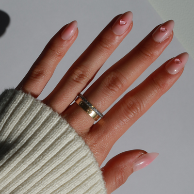 A close-up of a hand with long, almond-shaped nails painted using "Made You Blush Press-On Nails" by Glowie By Her, adorned with 3D gel heart accents. The hand is accessorized with a gold and a silver ring, each featuring small gemstones. Set against a plain, light surface background, the person is wearing a white ribbed sleeve.