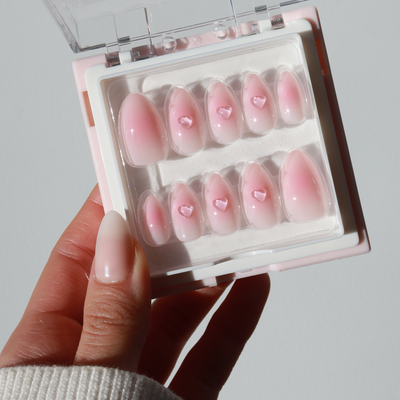 A hand with a manicured, pale pink nail holds a box of Glowie By Her's "Made You Blush Press-On Nails." These salon-quality faux nails feature a blush base with gradient pink hues and 3D gel heart accents on selected nails. The box has a transparent flip-top lid, neatly displaying the nails in two rows inside.