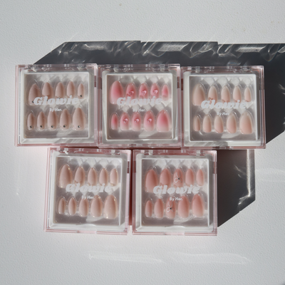 Five boxes from the "Sweet Talk Collection" of various styles and shades with a glossy finish, featuring baby pink press-on nails, are arranged on a white surface. Each box has a clear cover displaying the brand name "Glowie By Her." The shadows cast by the boxes suggest a sunny environment.