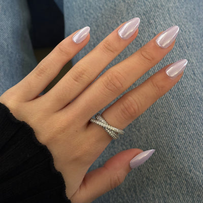 A hand adorned with Glowie By Her's Lavendare Press-On Nails, which are light purple and almond-shaped with salon-quality finish, rests on a denim background. The person is wearing a silver crisscross ring on their middle finger, while the sleeve of a black sweater partially covers their wrist.