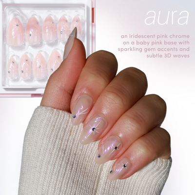 A hand adorned with Glowie By Her's Aura Press-On Nails, showcasing long, iridescent pink chrome nails with sparkling gem accents and subtle 3D wave patterns. The packaging for this reusable nail kit can be seen in the background. Text reads: "Aura - an iridescent pink chrome on a baby pink base with sparkling gem accents and subtle 3D waves.