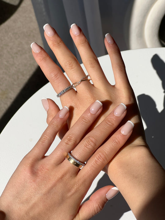 Hands with a salon-quality French manicure featuring Glowie By Her's Christi nails rest on a white surface, adorned with rings. Sunlight casts shadows, highlighting the customizable glossy finish of the Christi nails and textured bands. The background features a blurred white structure.