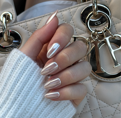 A hand adorned with long, glossy, salon-quality Allure Press-On Nails by Glowie By Her rests against a quilted, white handbag with shiny metal accents and dangling letter charms. The person is wearing a white knitted sweater.