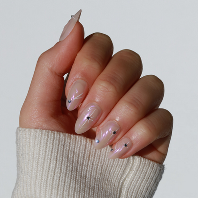 A hand adorned with long, glossy almond Aura Press-On Nails from Glowie By Her is shown. The nails feature a translucent finish embellished with tiny black star designs, each centered with small rhinestones. The hand is also wearing a cream-colored, ribbed sweater.