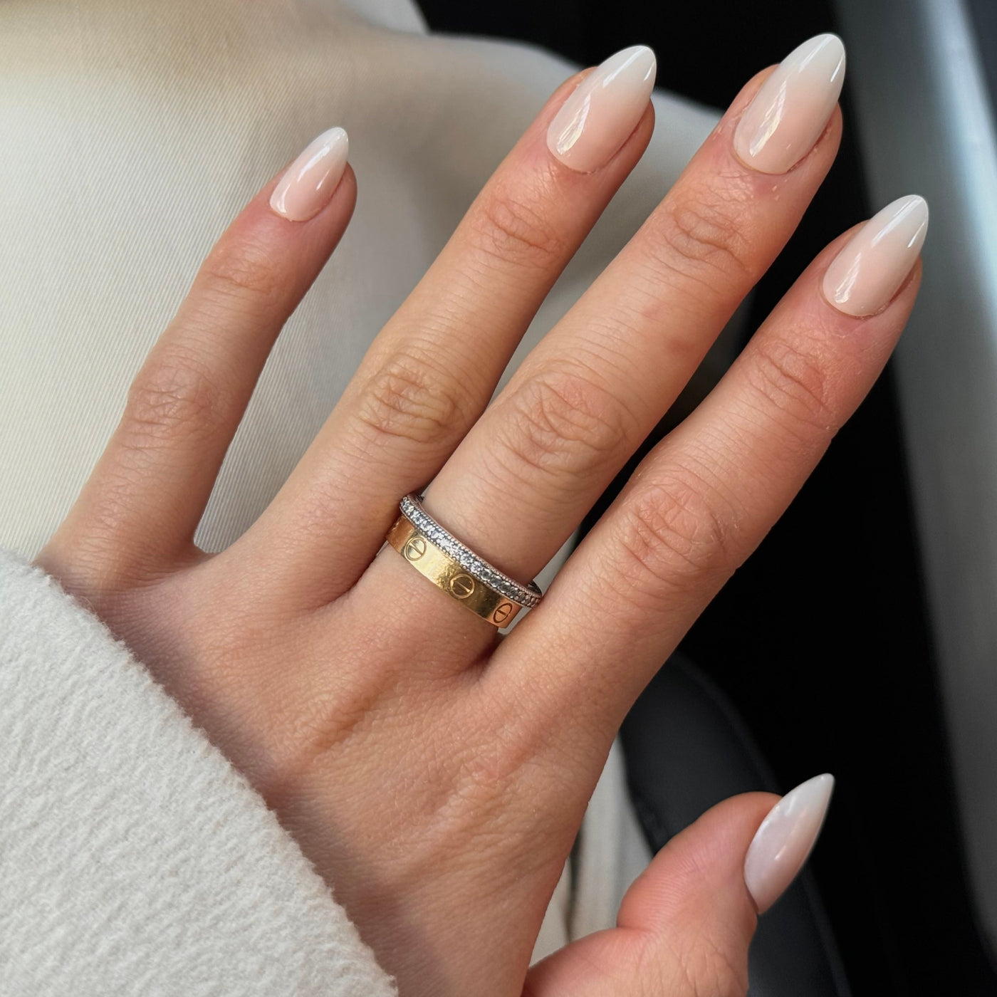 A hand displaying "Daydream Press-On Nails (Short)" by Glowie By Her in a milky ombre almond style, with two rings on the ring finger. One gold ring features engraved letters and symbols, while the other is a silver band adorned with small diamonds. The person is dressed in light-colored, soft fabric.