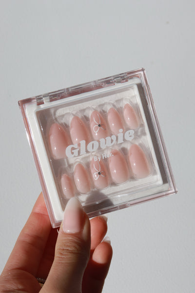 A hand holding an open plastic case containing a set of glossy, baby pink press-on nails labeled "Sweet Talk Collection by Glowie By Her (20% off)." The almond-shaped nails are arranged in rows within the case, and the person's nails are also painted in a light pink shade.