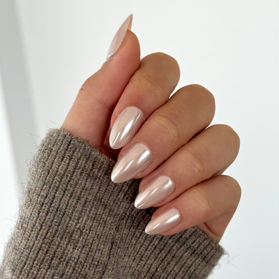 A close-up of a hand adorned with Glowie By Her's Allure Press-On Nails, featuring long, almond-shaped, salon-quality nails painted in a glossy, metallic light pink color. The hand is partially covered by the sleeve of a gray, ribbed sweater against a plain white background.