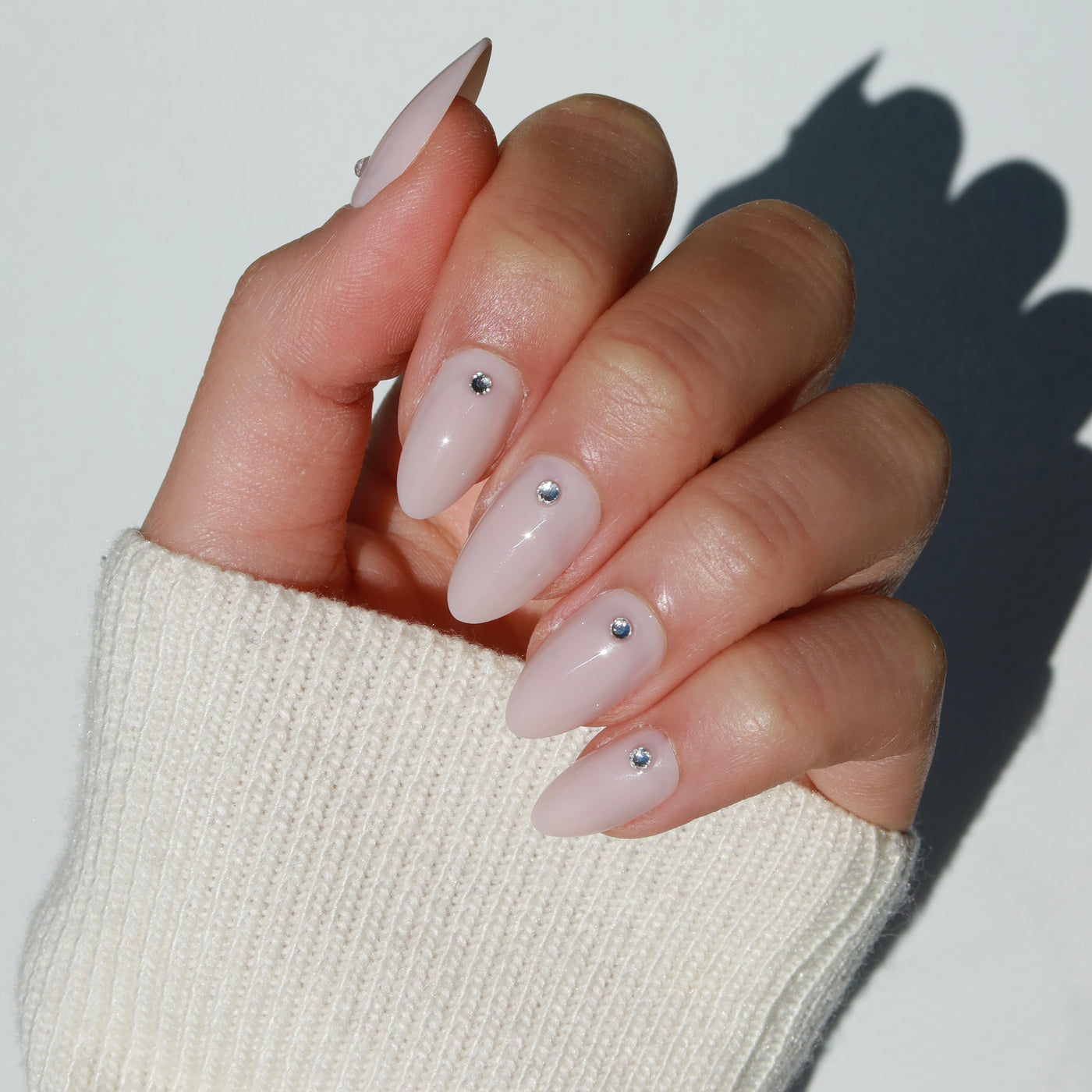 A hand with almond-shaped, baby pink Laura Press-On Nails by Glowie By Her, each adorned with a small glossy rhinestone near the cuticles, is shown against a plain background. The wearer is dressed in a cream-colored knitted sweater, visible in the lower portion of the image. These salon-quality nails add an elegant touch.