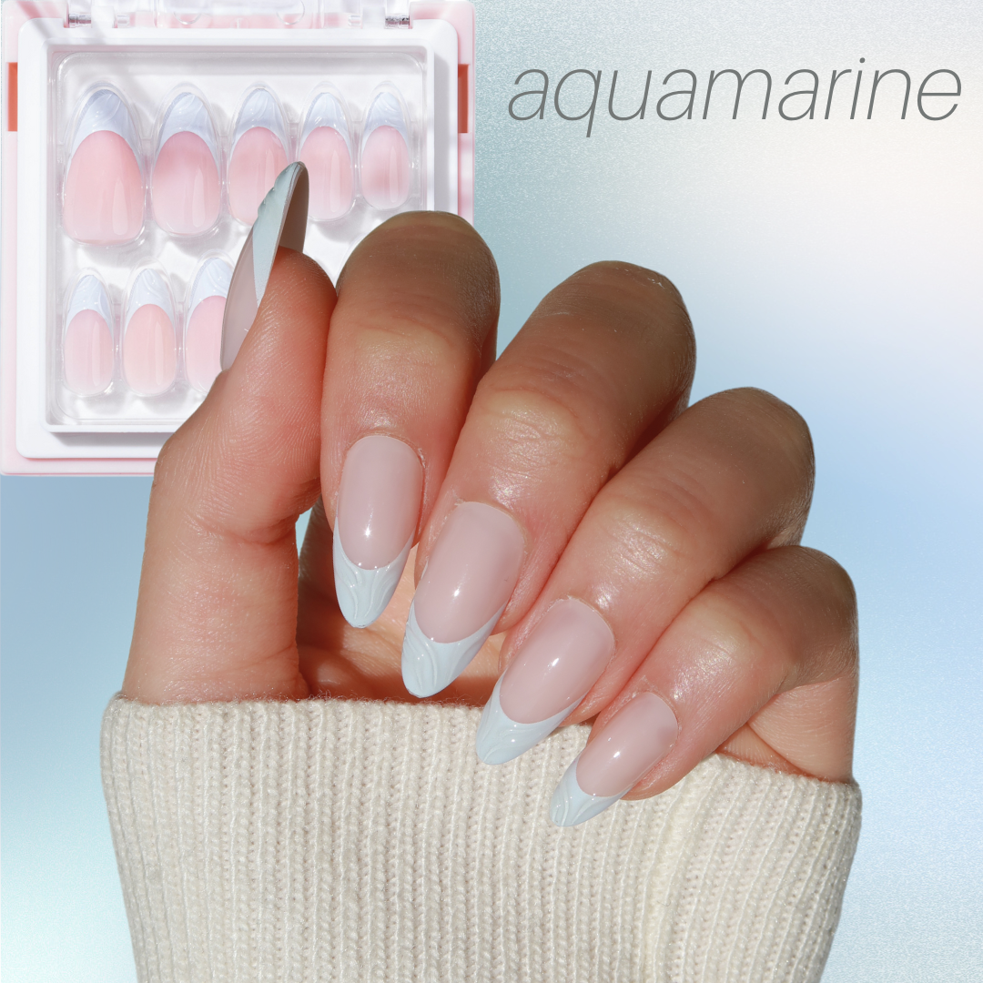 A hand featuring long, almond-shaped nails painted in a soft pink hue and decorated with light blue marble tips exemplifies the sophistication of Glowie By Her's Blue Mood collection. The individual is dressed in a cream sweater, while a package of these salon-quality nails can be seen in the background, marked with "aquamarine" in the corner and labeled as Blue Mood (NEW - 25% Off).