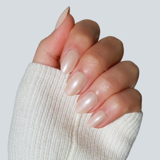 A hand with salon-quality, almond-shaped nails painted in the glossy, pale nude shade of Glowie By Her's Short & Sweet (10% Off) emerges gracefully from the sleeve of a white knitted sweater.