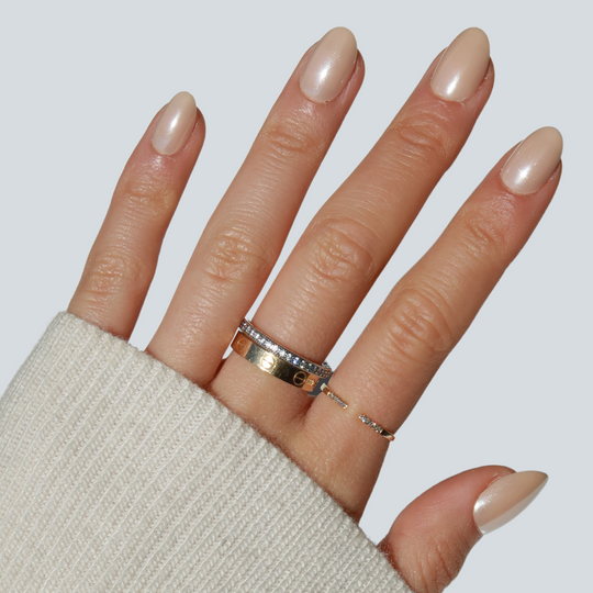 A hand with salon-quality manicured nails painted in "Bubbly" by Glowie By Her is adorned with three gold rings, one of which is embellished with small diamonds. The light beige sweater sleeve partly covers the wrist, adding an elegant touch to the chic ensemble.