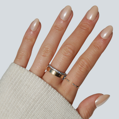 A hand adorned with a light beige sweater sleeve showcases beautifully manicured nails featuring Bubbly Press-On Nails from Glowie By Her. The nude, salon-quality finish is complemented by four gold rings, including one embedded with diamonds and an elegant band with small circular cutouts. This setup flawlessly captures the neutral coffee/beige chrome style against a light gray background.