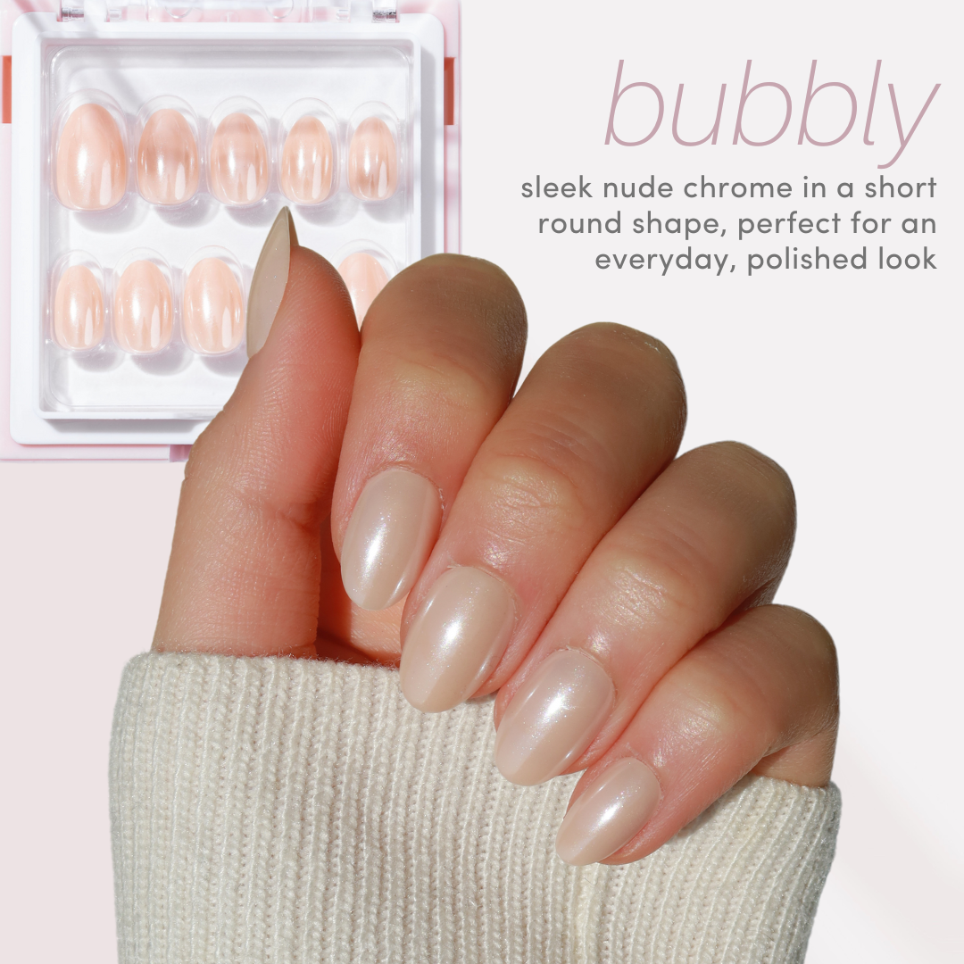 A hand with sleek nude chrome, short round-shaped nails holds a cream sweater. In the background, a box from Glowie By Her’s Chrome Crush (10% Off) collection is displayed. Text reads: "bubbly - perfect for an everyday, polished look.