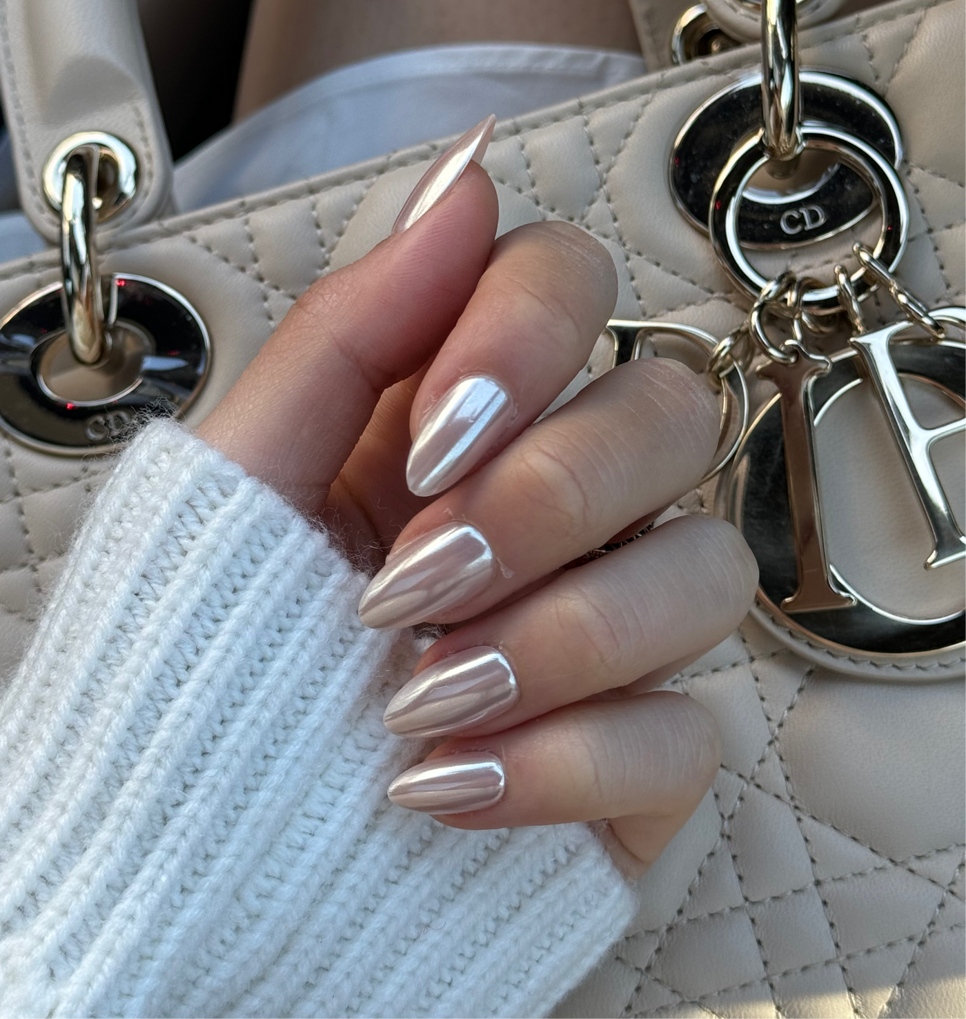 A close-up image of a hand with long, glossy, chrome Allure Press-On Nails in an almond shape by Glowie By Her. The hand is wearing a white ribbed knit sleeve and is holding a beige quilted handbag adorned with silver chains and a keychain featuring the letters "CD." These salon-quality press-on nails add an extra touch of elegance.