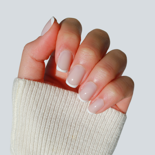 A hand with a salon-quality manicure featuring Glowie By Her's Christi pale pink and customizable white French tips is shown against a light background, partially covered by a cream-colored sweater sleeve.