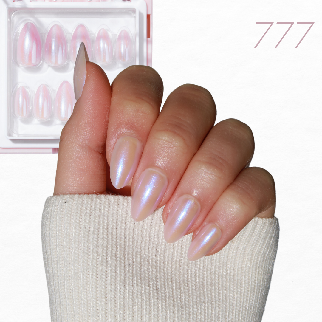 A hand adorned with iridescent pink, almond-shaped nails from Glowie By Her's Chrome Crush Collection peeks through a light beige sleeve. In the background, there's a set of similar Cosmic Dreams vegan & cruelty-free false nails displayed in a clear box, featuring the number "777" written in pink.