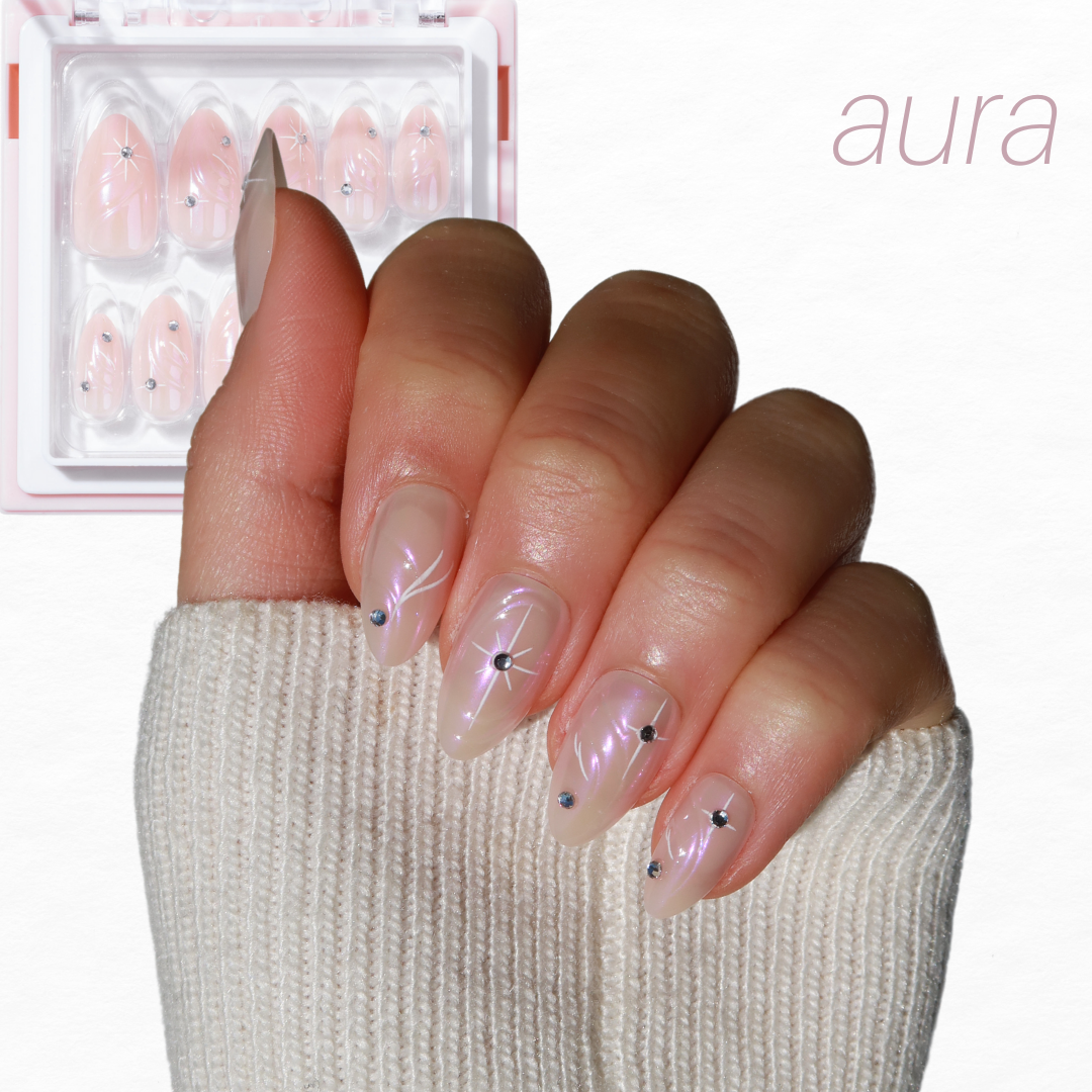 A hand with long, almond-shaped nails showcases an iridescent design featuring small star and dot accents. The glossy pink nails highlight the Cosmic Dreams set from the Chrome Crush Collection by Glowie By Her, now available at 30% off. Delicate nail art shines through a soft beige sleeve partially covering the hand, with the word "aura" elegantly inscribed in the top right corner.