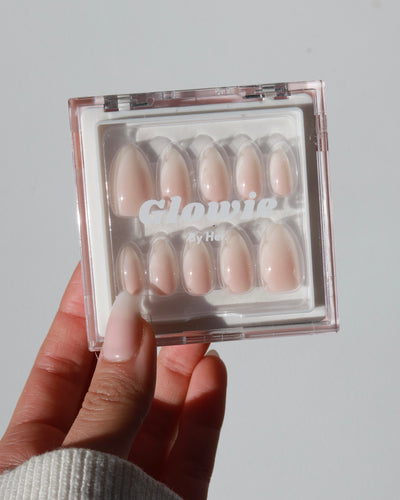 A hand holding a clear plastic case with "Glowie By Her" printed on it, showcasing the Sweet Talk Collection (20% off). Inside the case are ten pale pink artificial nails with a glossy finish, neatly arranged in rows. The person holding the case is wearing a white, ribbed long-sleeve shirt and has light pink painted nails.