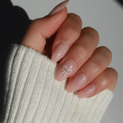 A hand adorned with long, almond-shaped baby pink Cutie Press-On Nails from Glowie By Her showcases a delicate white bow design with a small gem at the center. The customizable nails are partially covered by the sleeve of a cream-colored, ribbed sweater.